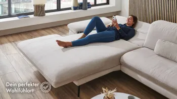 Frau liegt entspannt auf ausziehbarer heller Stoffcouch in modernem Wohnzimmer mit großen Fenstern und Holzfußboden – zeigt den Komfort und die Funktionalität der Couch als gemütlichen Rückzugsort.